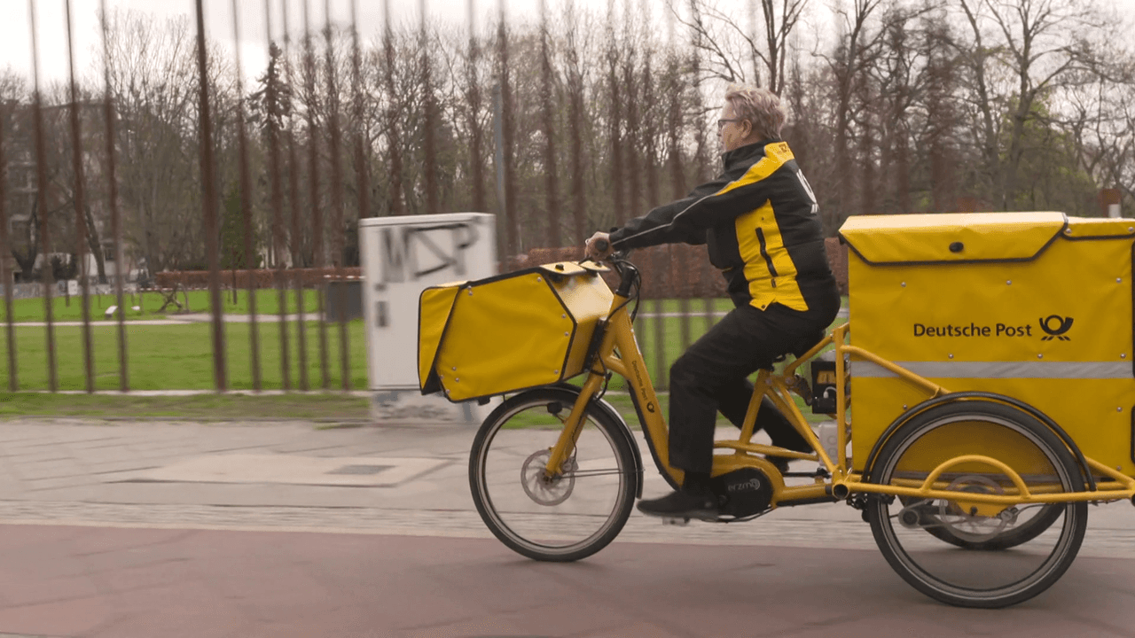 Eine Postbotin transportiert Lieferungen auf einem Fahrrad. Screenshot aus Presse-Video aus der DHL-Mediathek (erstellt von Laura Wenzel)