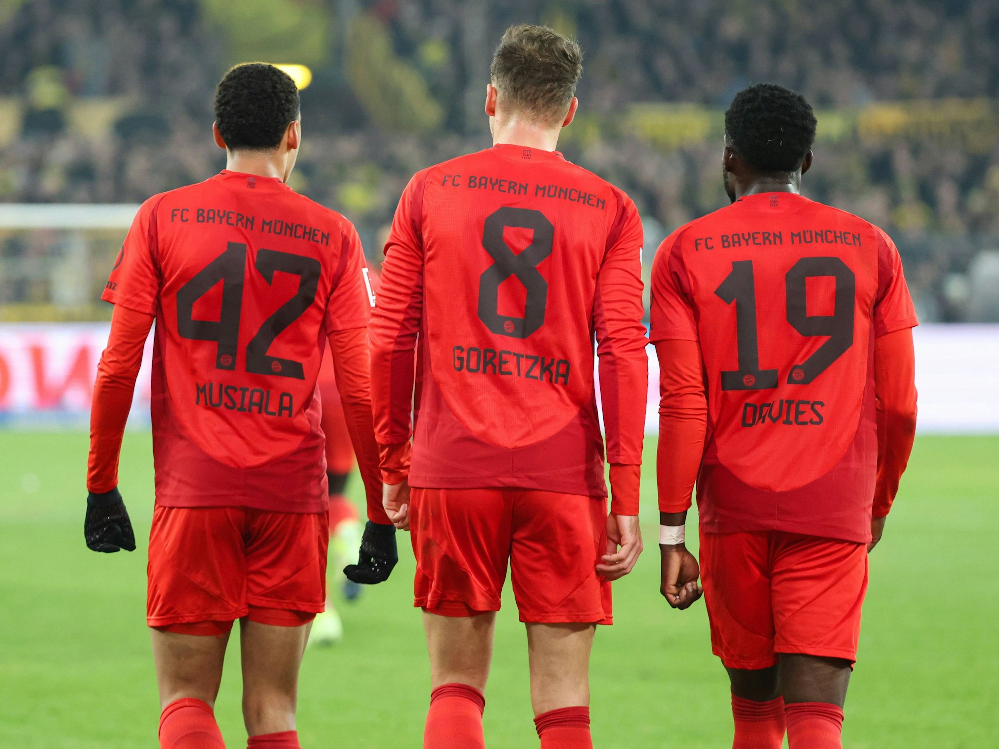 Jamal Musiala, Leon Goretzka und Alphonso Davies gehen nebeneinander.