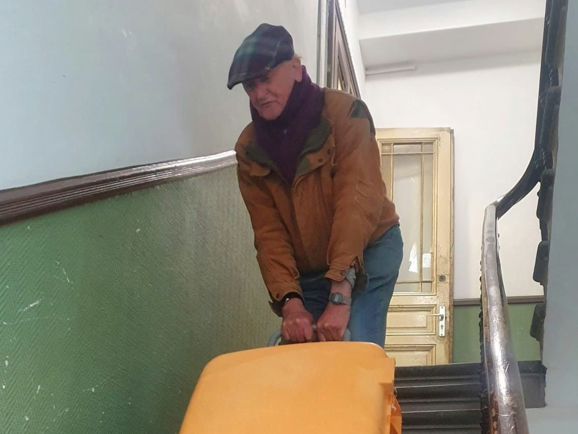 Ein alter Herr bringt einen Koffer die Treppen eines Treppenhauses hinunter.