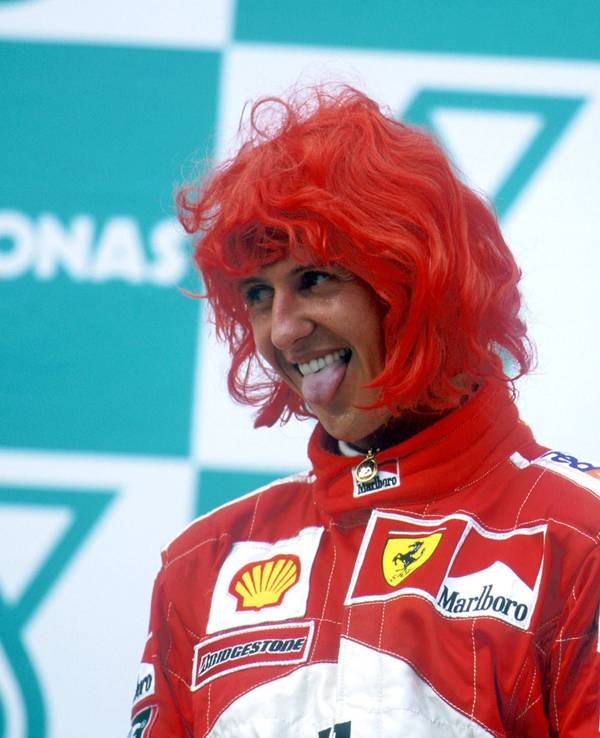 Michael Schumacher steht mit roter Perücke auf dem Siegertreppchen und streckt die Zunge raus.