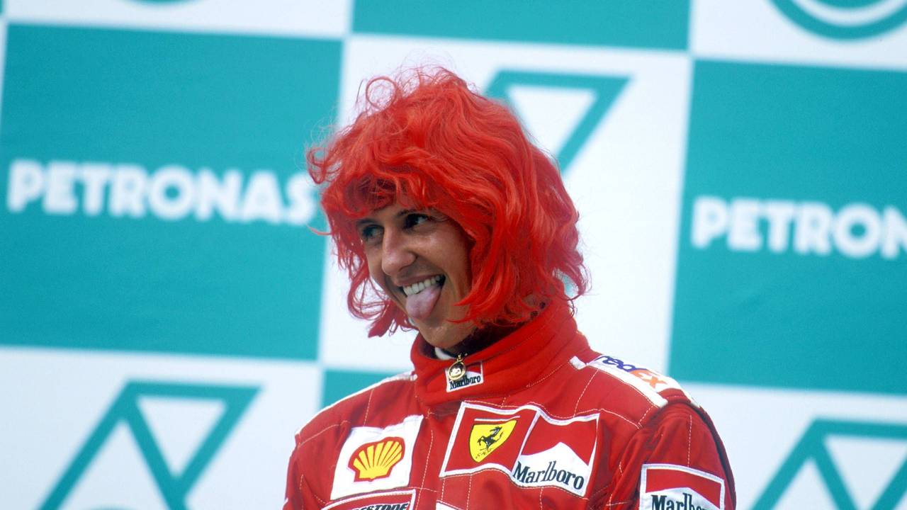 Michael Schumacher steht mit roter Perücke auf dem Siegertreppchen und streckt die Zunge raus.