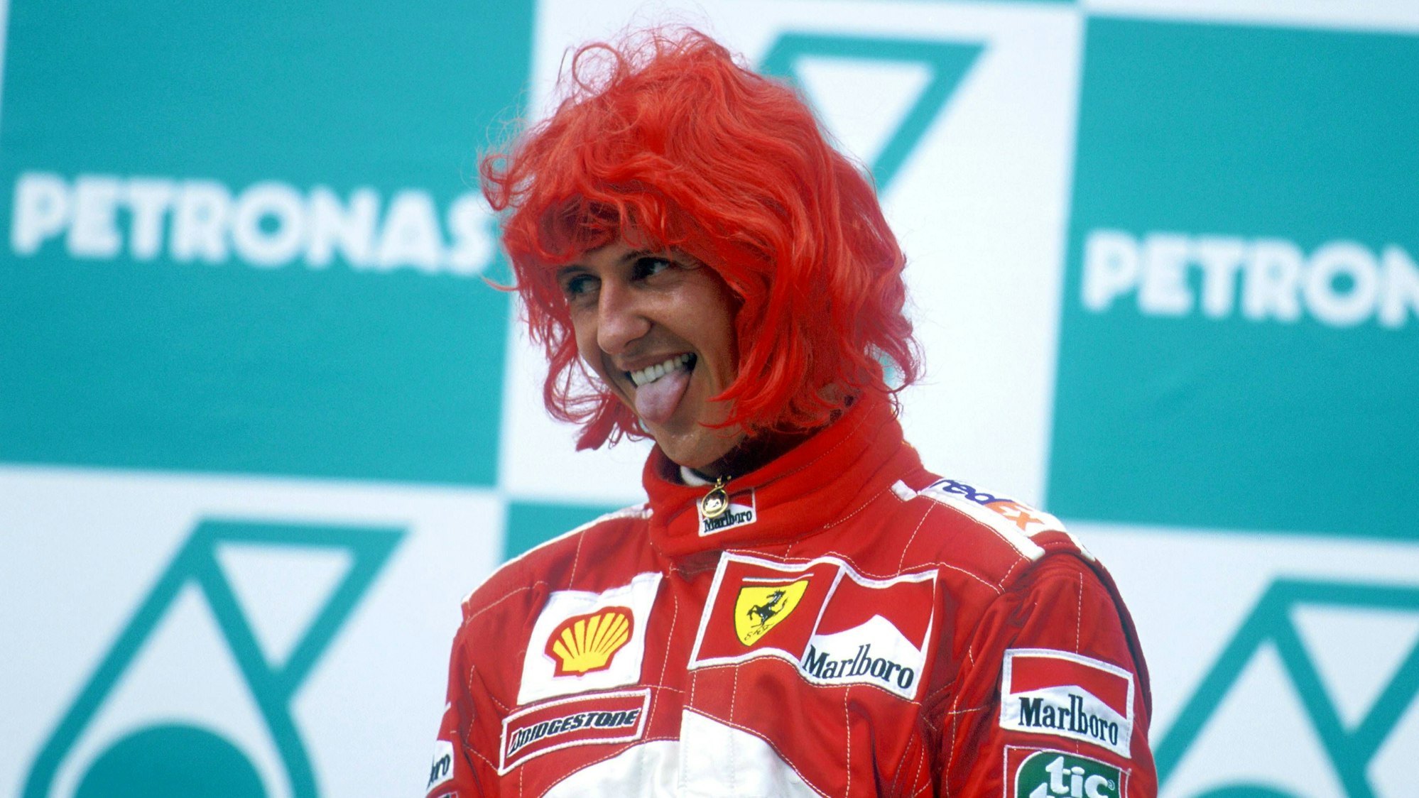 Michael Schumacher steht mit roter Perücke auf dem Siegertreppchen und streckt die Zunge raus.