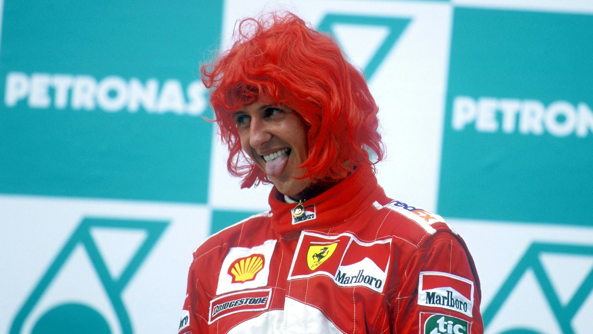 Michael Schumacher steht mit roter Perücke auf dem Siegertreppchen und streckt die Zunge raus.
