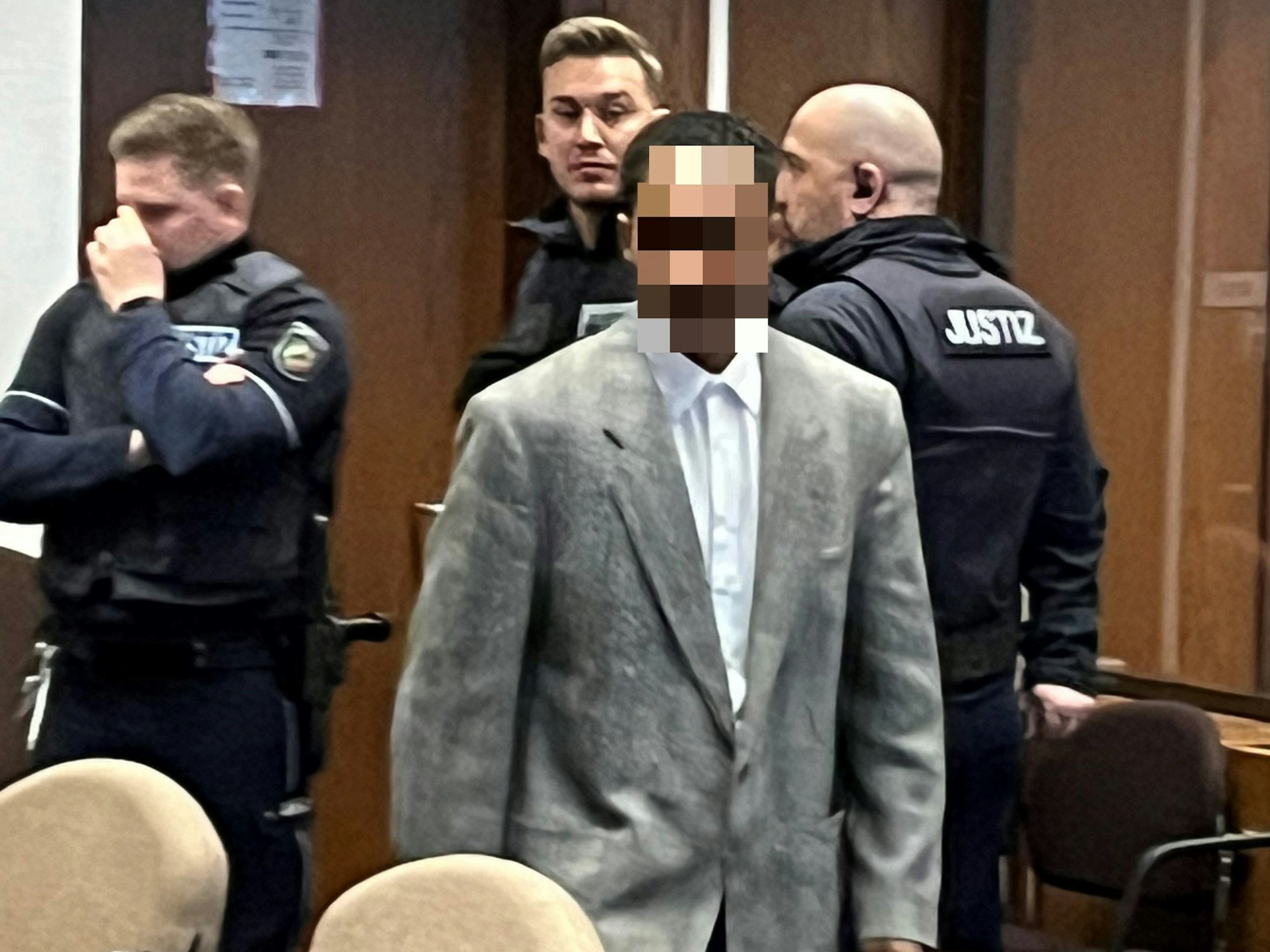 Ein junger Mann, der ein Jackett trägt, geht in einem Gerichtssaal an drei Justizwachtmeistern vorbei.