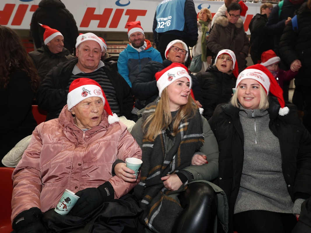 Zwei junge Frauen singen mit einer älteren Dame, alle drei tragen Weihnachtsmannmützen.