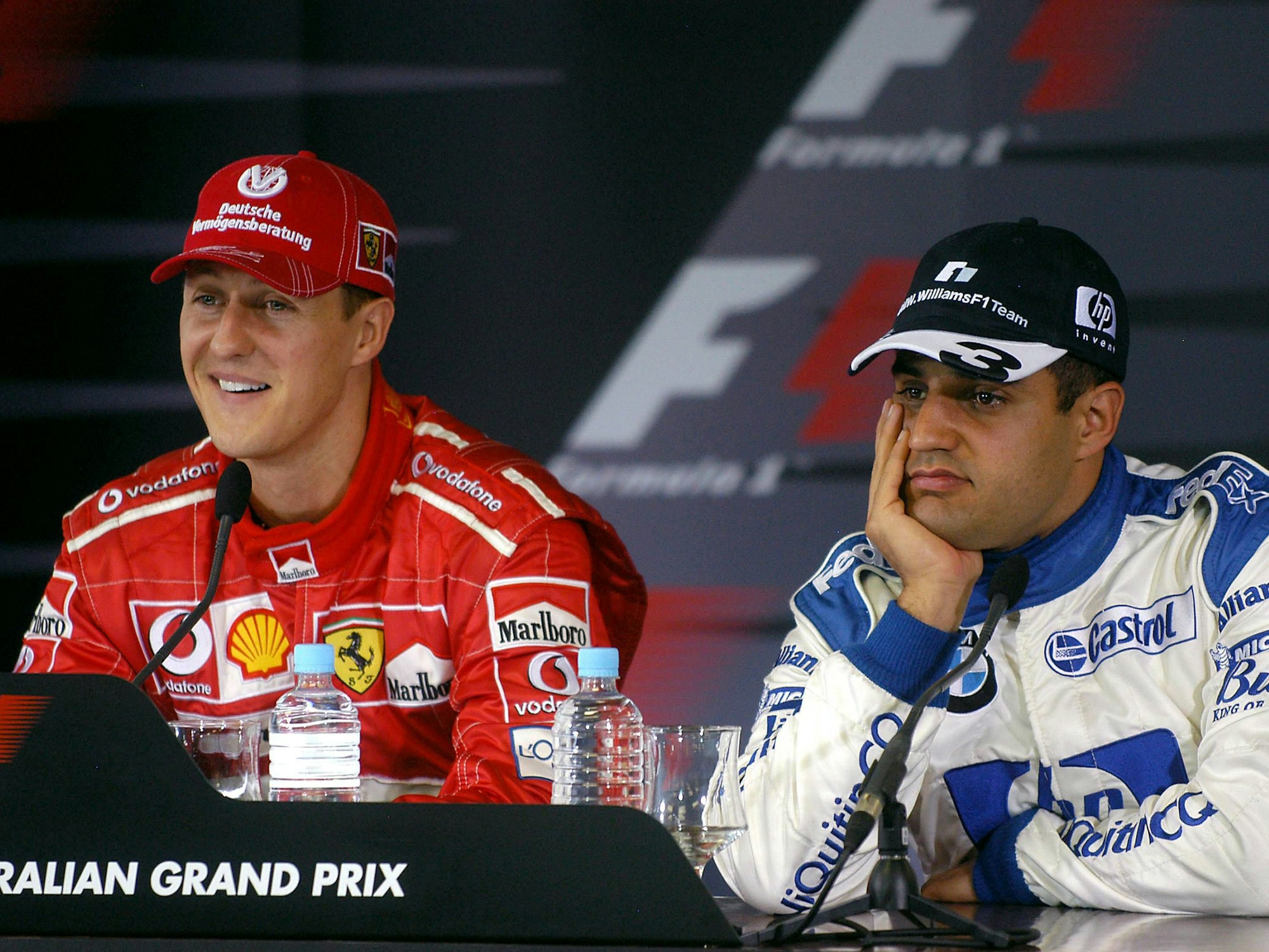 Michael Schumacher lächelt bei einer Pressekonferenz, Juan Pablo Montoya sietzt gelangweilt daneben.