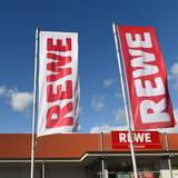 Fahnen mit dem Logo des Lebensmitteleinzelhändlers Rewe wehen vor einer Filiale.