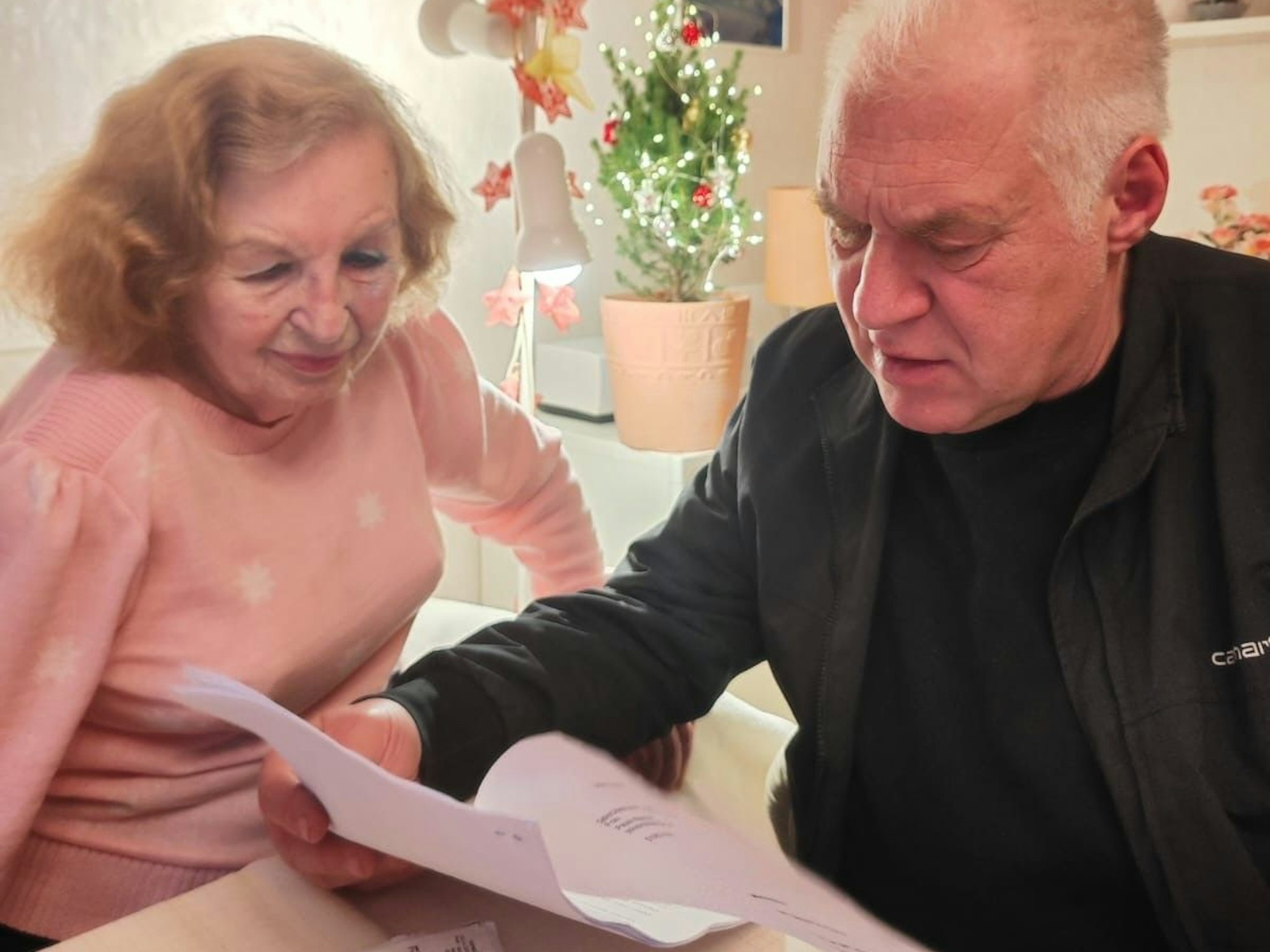 Ein Mann guckt auf ein Schreiben in seiner Hand, neben ihm am Tisch sitzt eine alte Dame