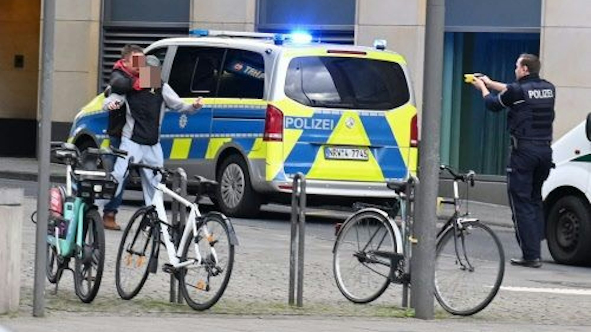 Ein Mann hält einen anderen vor sich, ein paar Meter weiter steht ein Polizist und richtet einen Taser auf ihn.