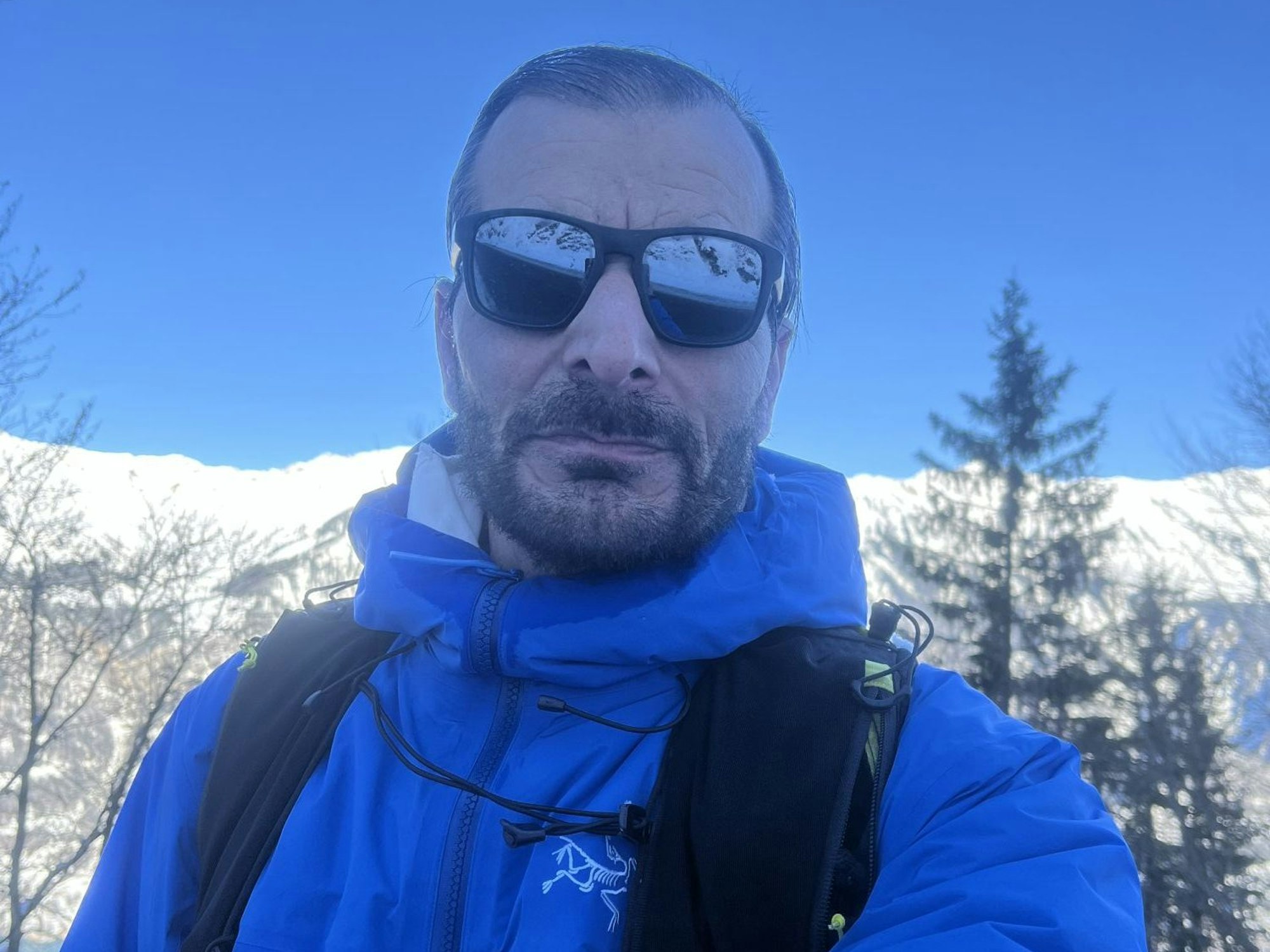 Ein Mann mit Sonnenbrille schaut in die Kamera. Im Hintergrund sind schneebedeckte Berge.
