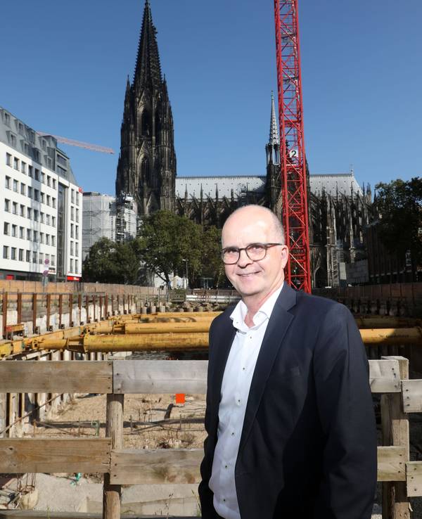 Markus Greitemann steht vor einer großen Baustelle in Köln, im Hintergrund ist der Dom zu sehen.