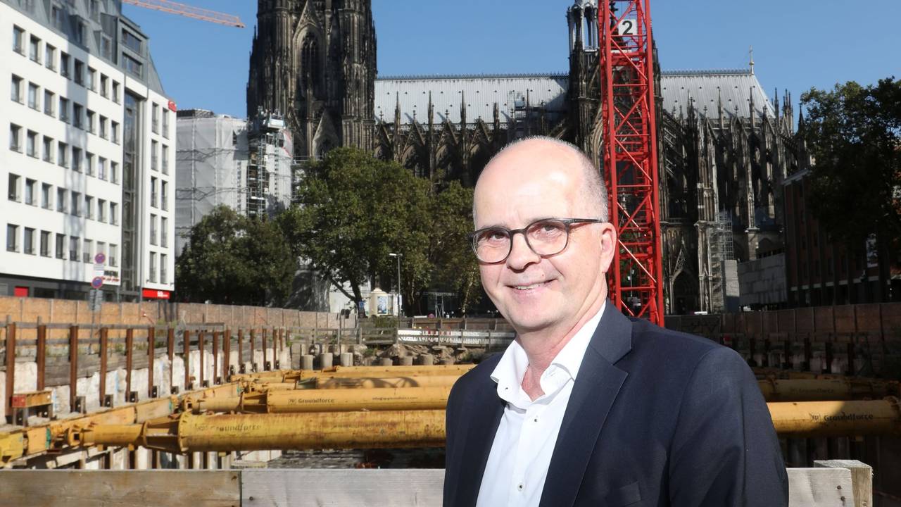 Markus Greitemann steht vor einer großen Baustelle in Köln, im Hintergrund ist der Dom zu sehen.