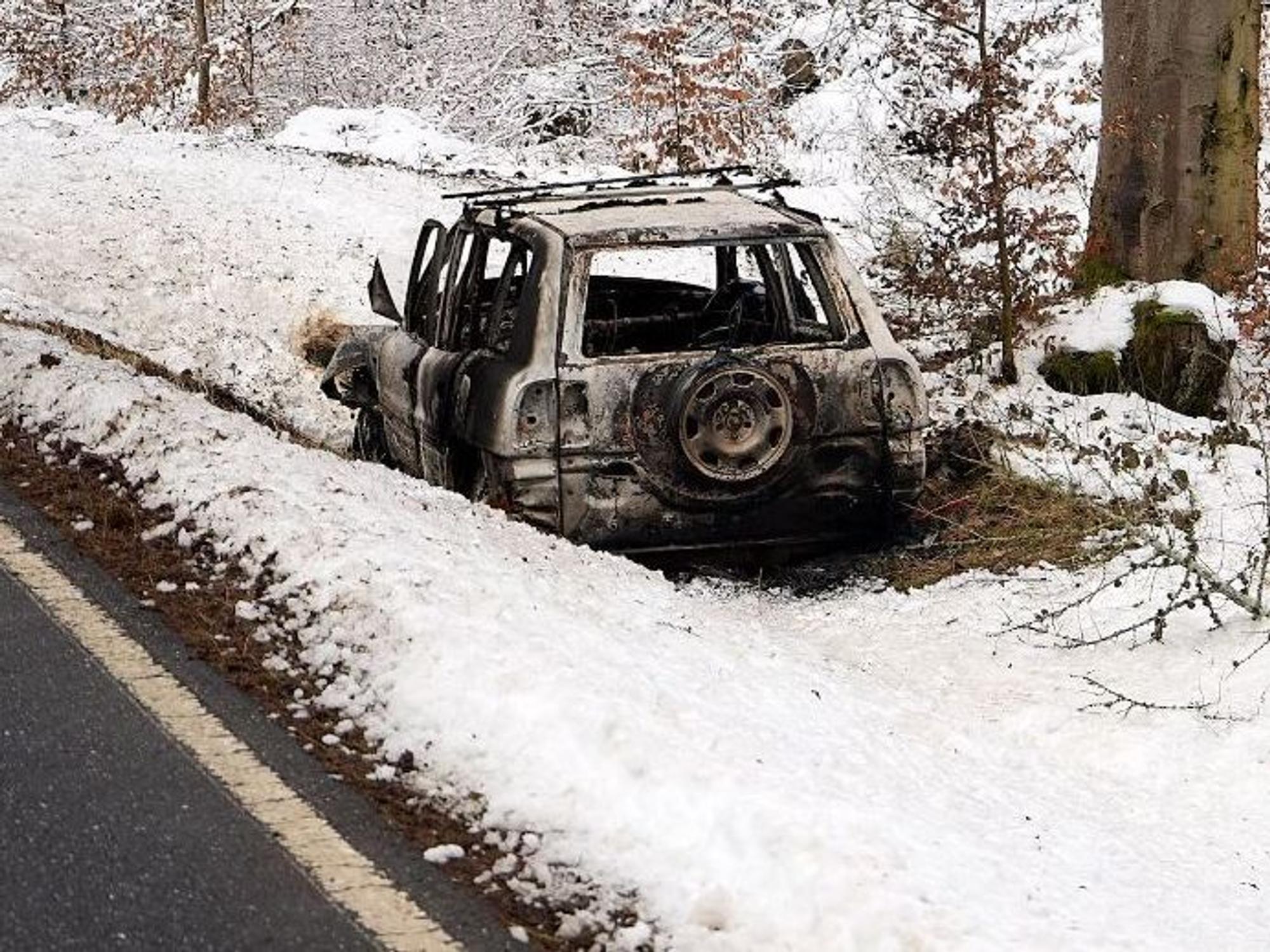 Ein ausgebranntes Fahrzeug steht im Schnee neben einer Straße.