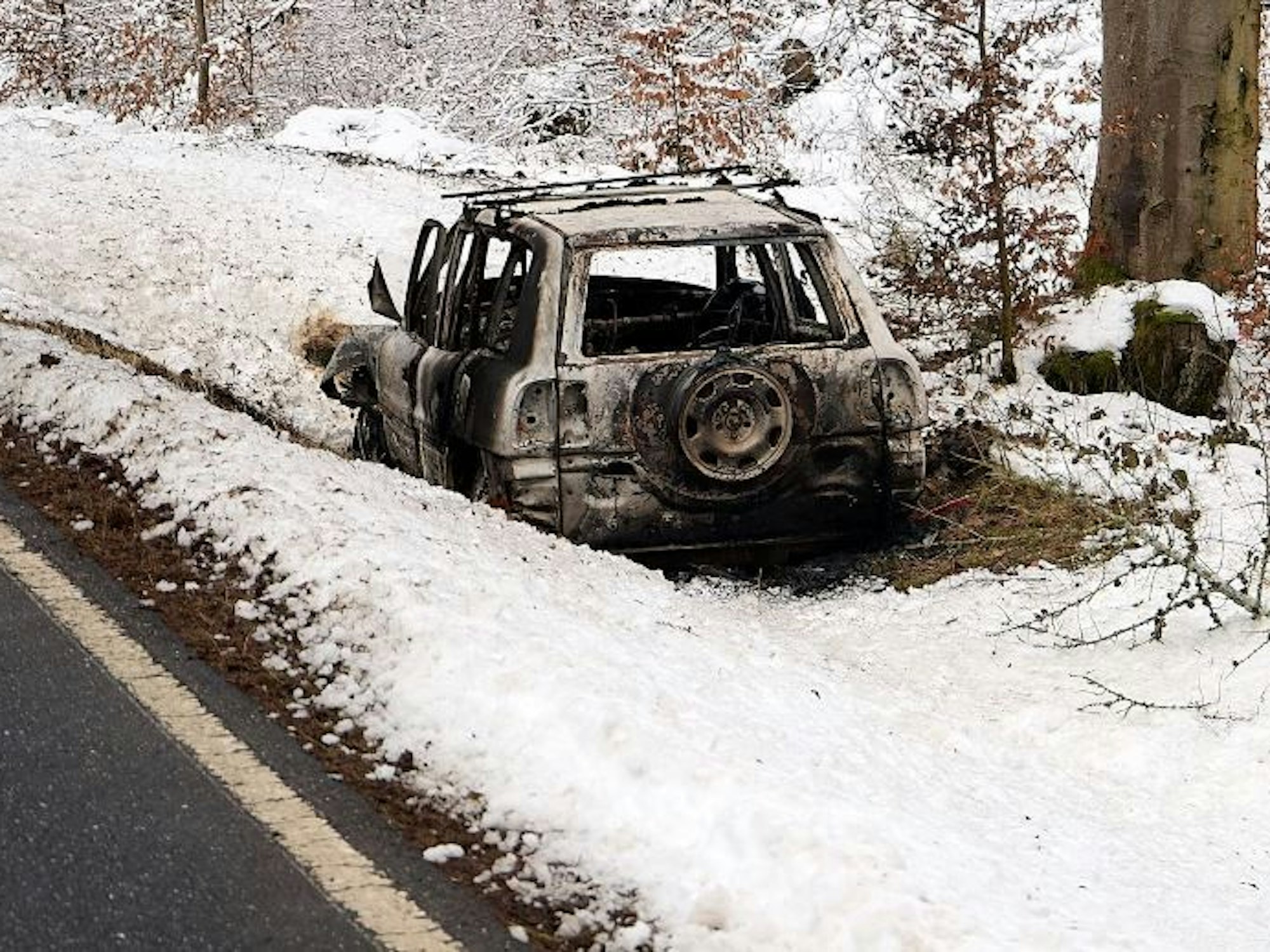 Ein ausgebranntes Fahrzeug steht im Schnee neben einer Straße.