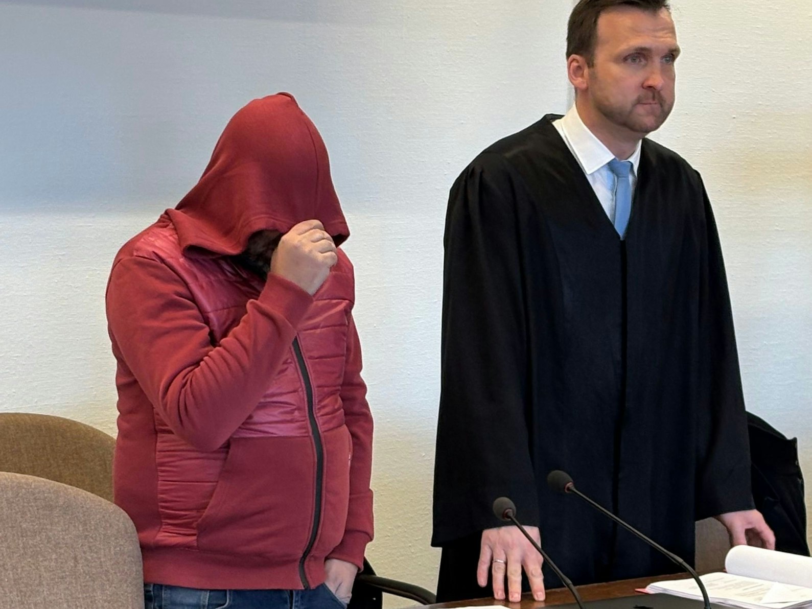 Ein Mann zieht die Kapuze seines Pullis bis tief ins Gesicht, neben ihm steht sein Anwalt.