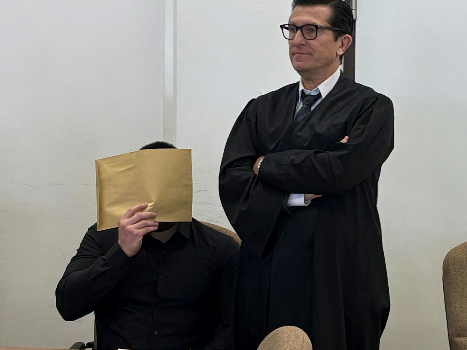 Ein sitzender Mann hält sich einen großen Briefumschlag vor das Gesicht, neben ihm steht sein Anwalt mit verschränkten Armen.