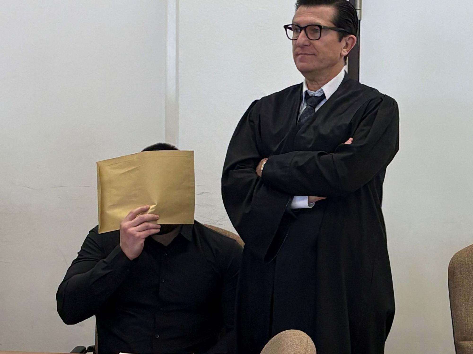 Ein sitzender Mann hält sich einen großen Briefumschlag vor das Gesicht, neben ihm steht sein Anwalt mit verschränkten Armen.