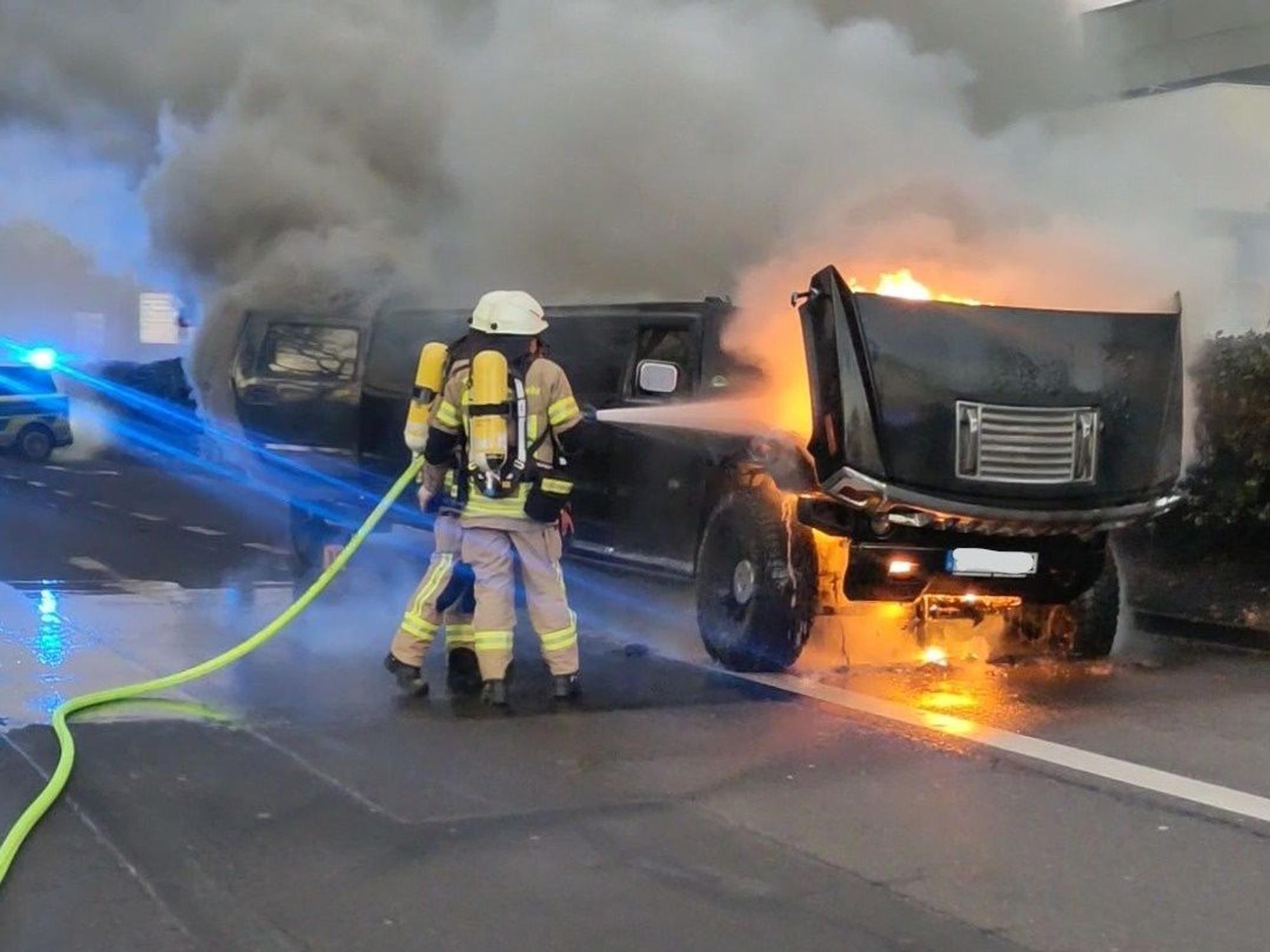 Feuerwehr steht an einem brennenden Auto, im Hintergrund steigt dichter Rauch auf.