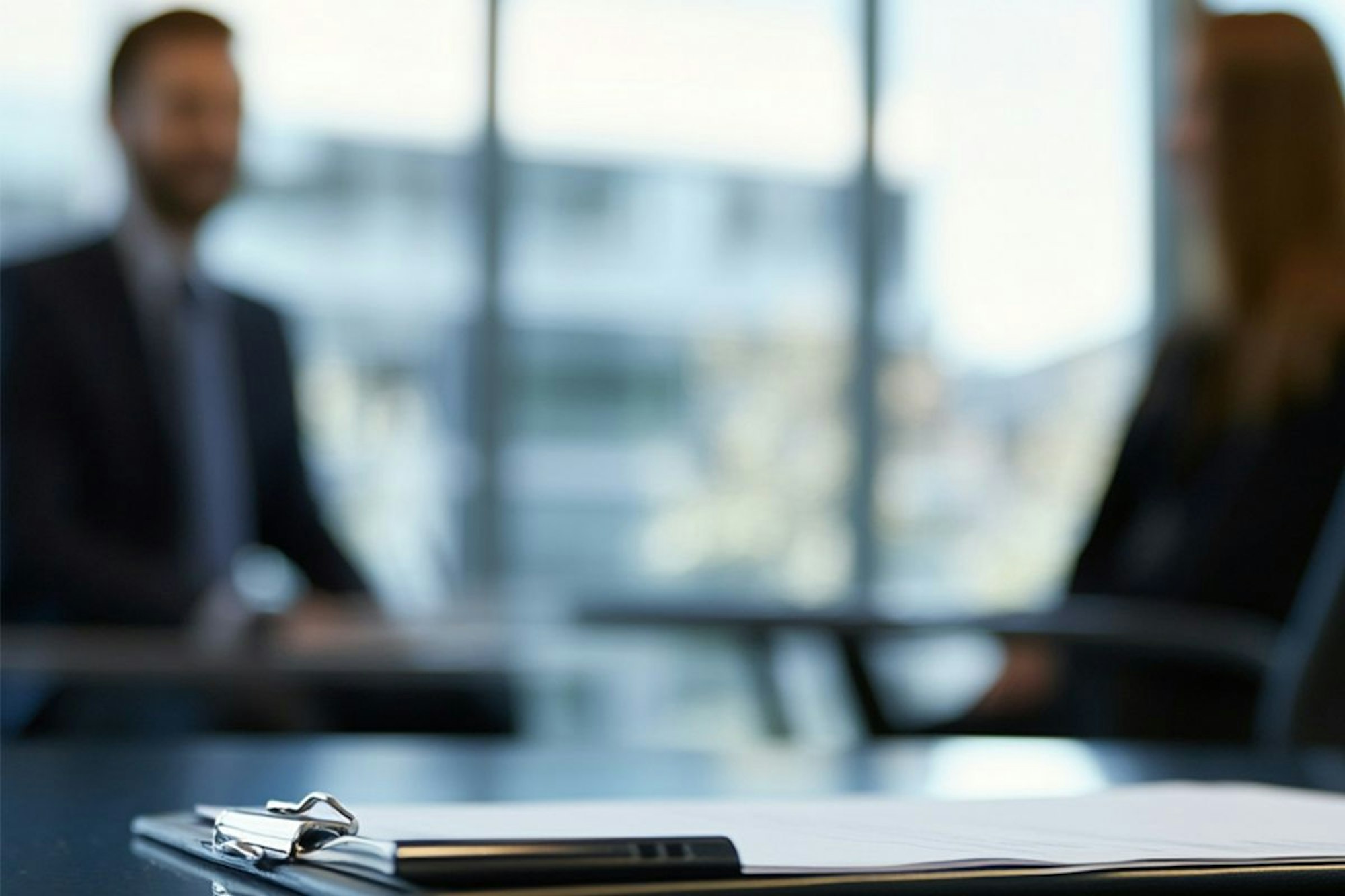 Das Bild zeigt zwei Personen in einem Büro unscharf im Hintergrund. Im Vordergrund liegt ein Klemmbrett mit Papier. Das Bild spiegelt eine Bewerbungssituation wider.