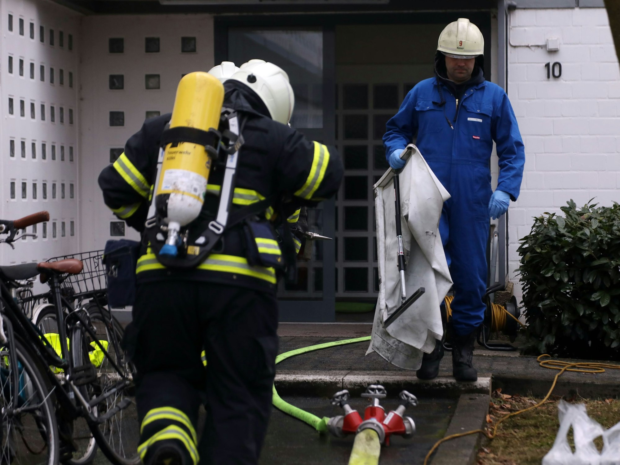 Zwei Feuerwehrleute tragen Atemschutz und gehen auf eine Haustüre zu, ihnen entgegen kommt ein Kollege, der einen Overall und einen Helm trägt.