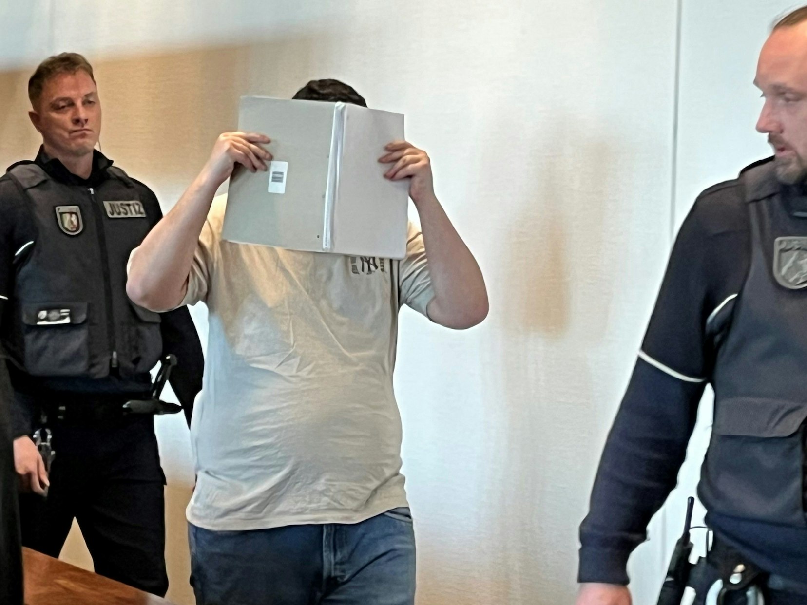 Zwei Justizwachtmeister begleiten einen dicklichen Mann, der sein Gesicht mit einem Schreibblock verdeckt, in den Gerichtssaal.