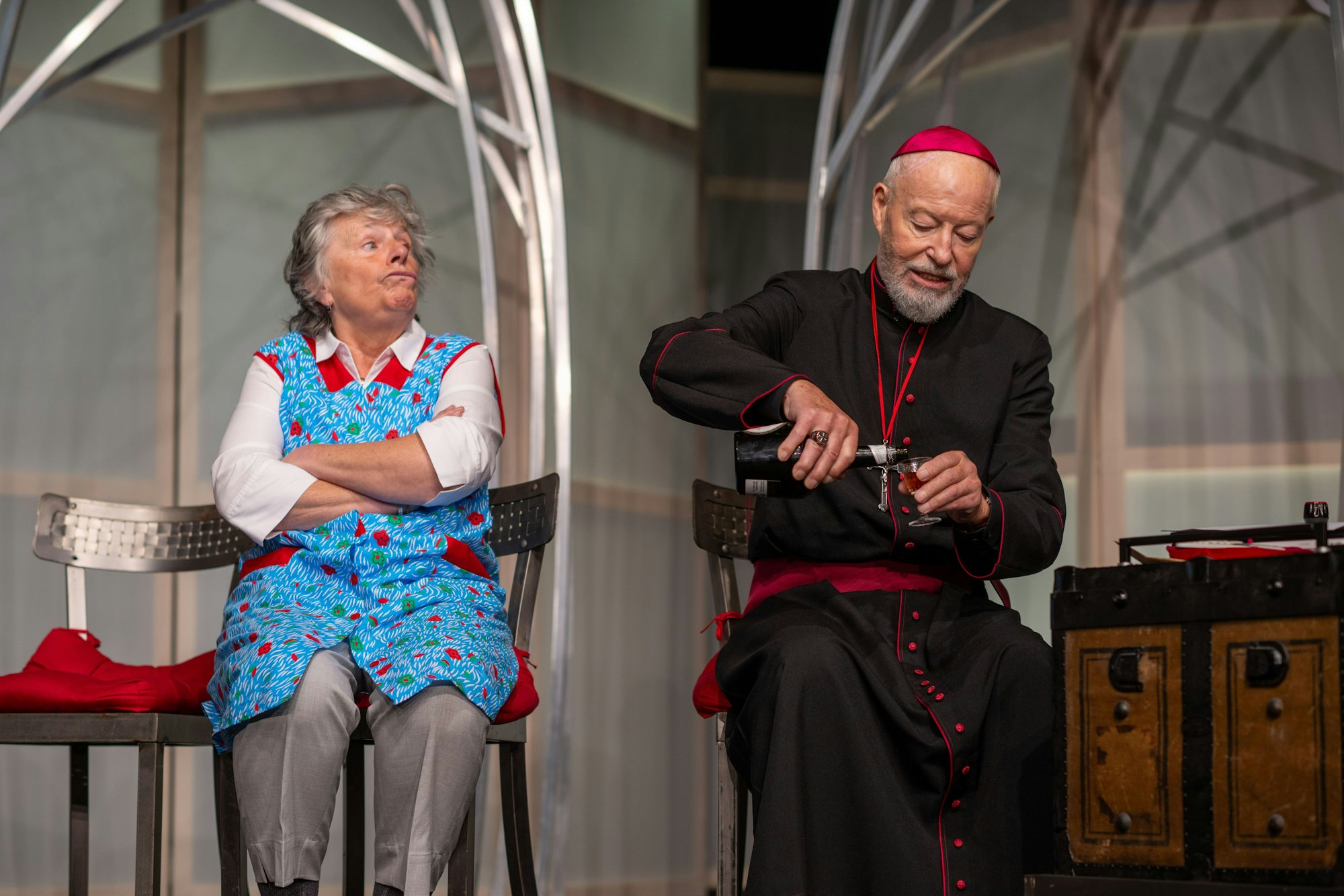 Ein als Kardinal verkleideter Mann gießt sich ein Getränk ein, daneben sitzt eine Frau im Kittel.