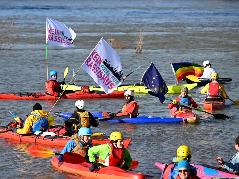 Menschen in Kanus sind auf dem Wasser und halten Fahnen mit der Aufschrift „Kein Rassismus“.