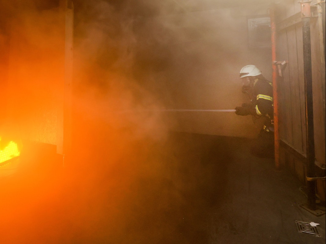 Ein Feuerwehr-Azubi hält mit einem Löschschlauch auf ein Feuer.