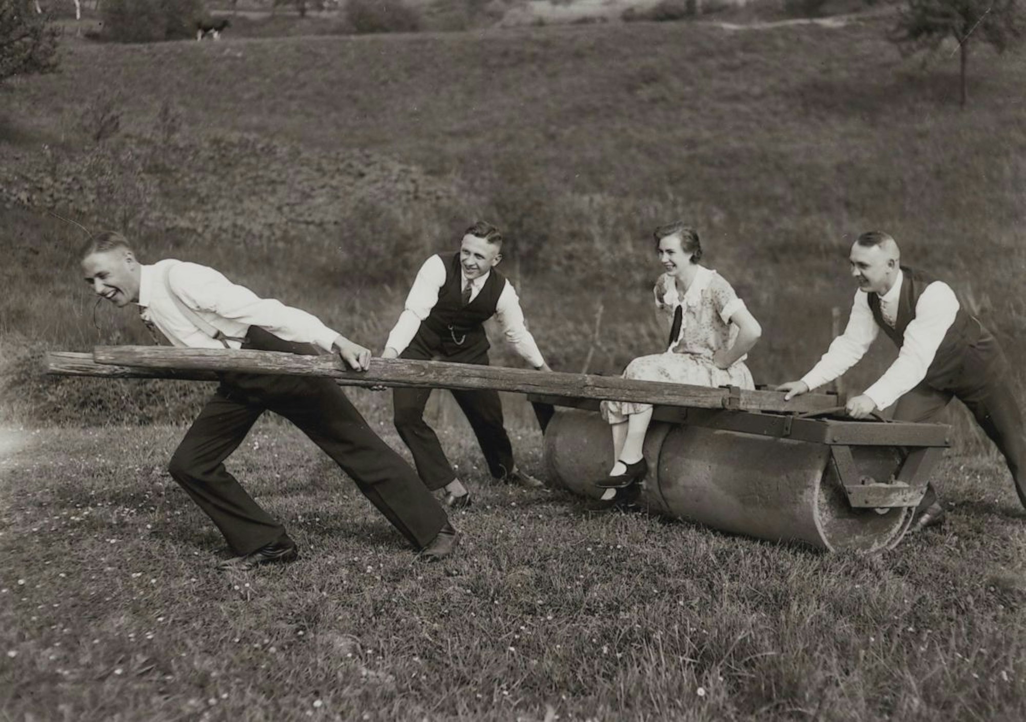Drei Männer ziehen einen alten Pflug, auf dem eine Frau sitzt. Altes Bild in Schwarz-Weiß.