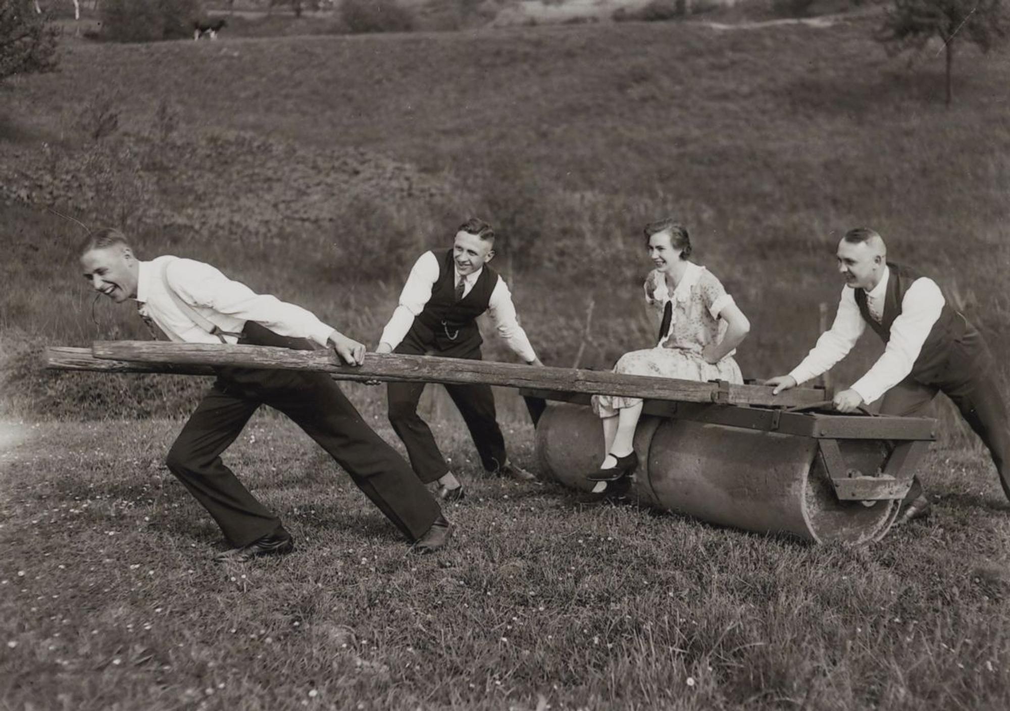 Drei Männer ziehen einen alten Pflug, auf dem eine Frau sitzt. Altes Bild in Schwarz-Weiß.