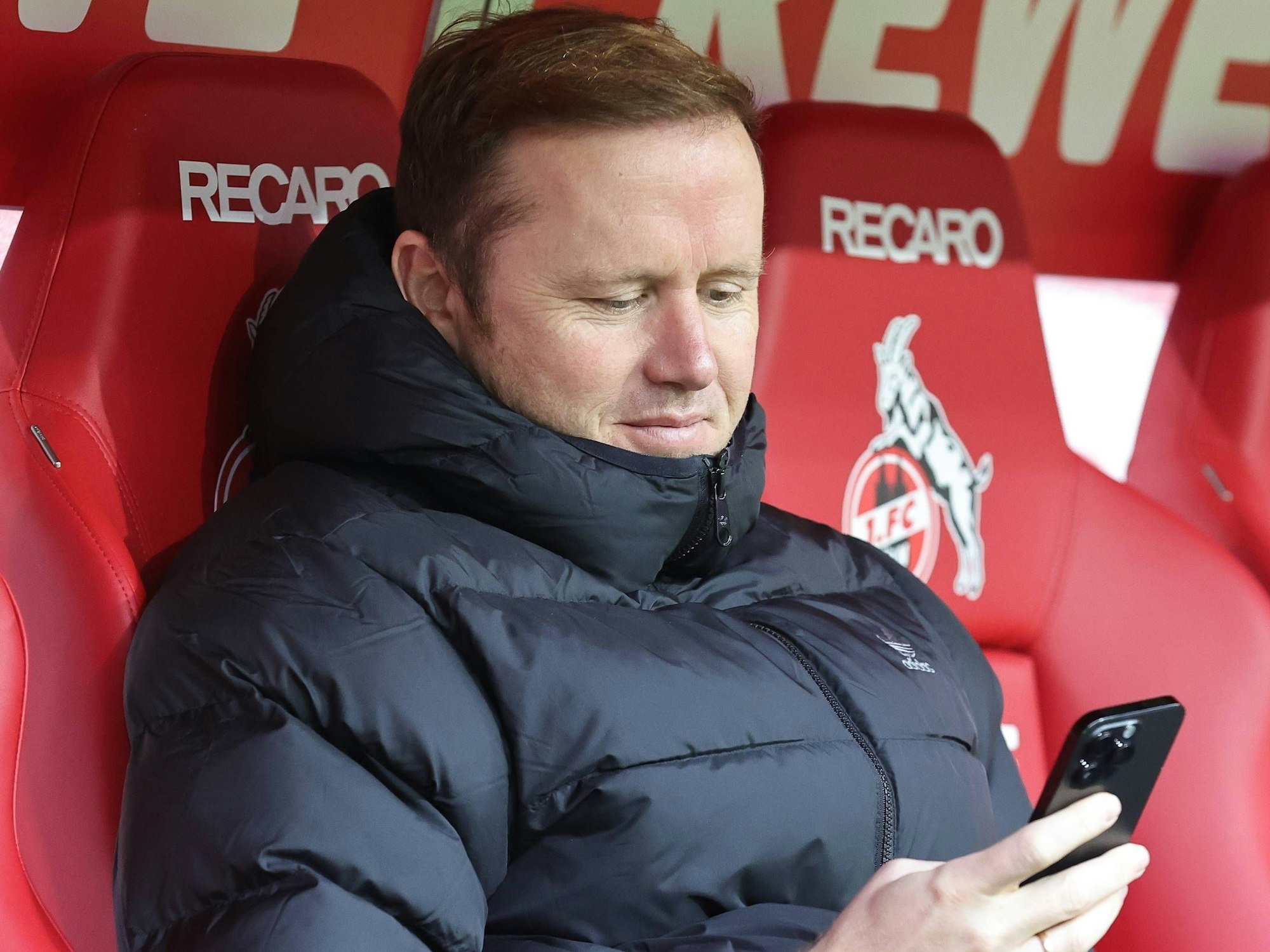 Olaf Rebbe sitzt auf der Bank im Rhein-Energie-Stadion mit seinem Handy in der Hand.