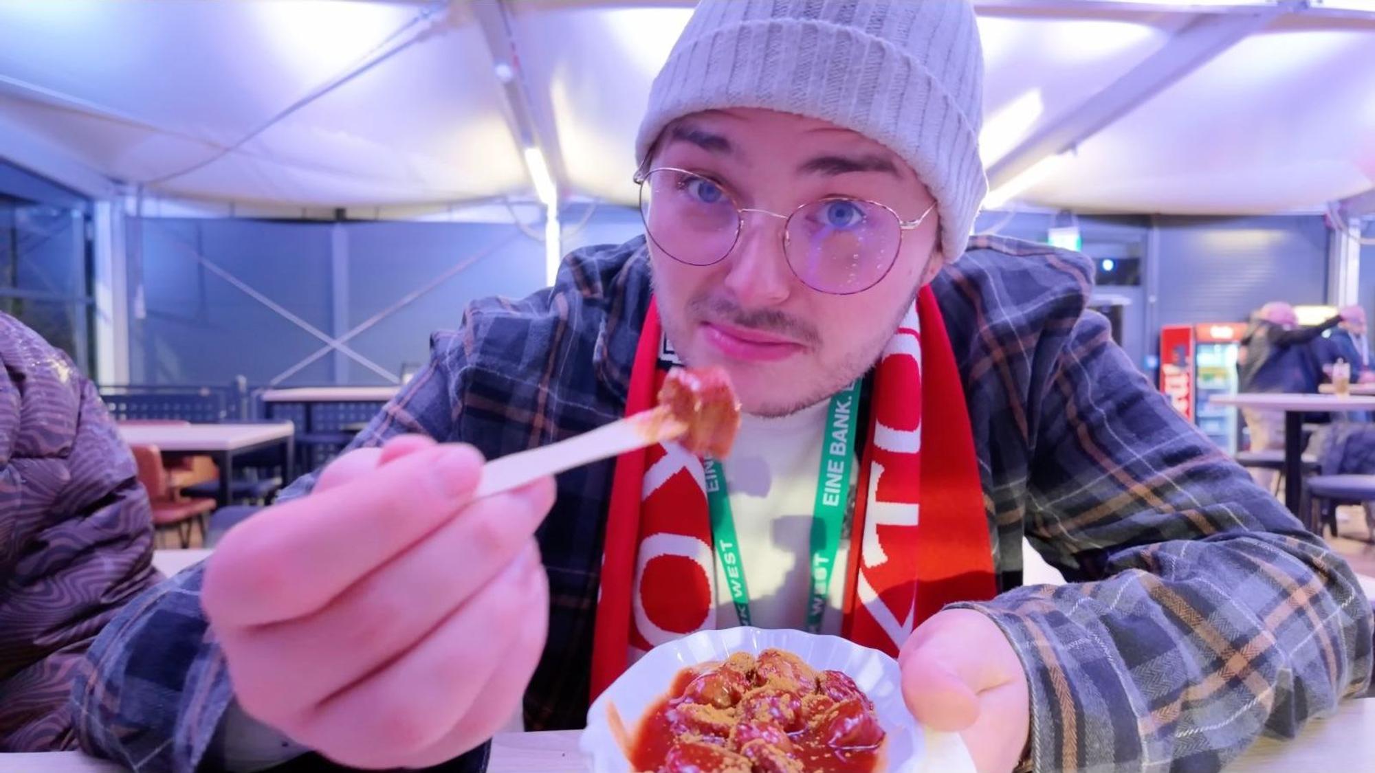 Der YouTuber Benji testet die Currywurst bei Viktoria Köln und guckt irritiert in die Kamera.