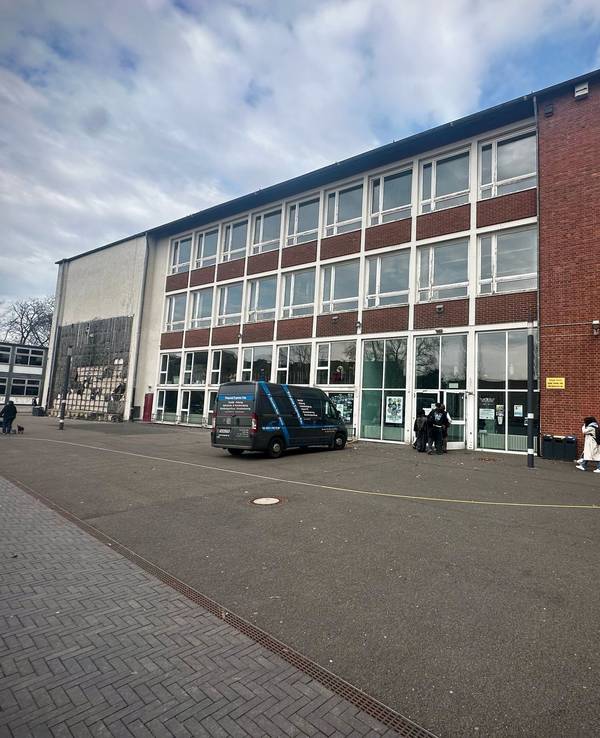 Mehrere Personen gehen auf den Eingang eines zweistöckigen Schulgebäudes zu. Davor steht ein Fahrzeug.