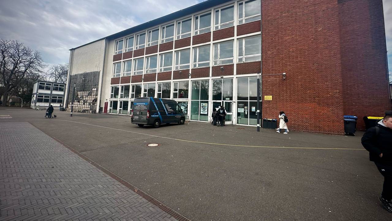 Mehrere Personen gehen auf den Eingang eines zweistöckigen Schulgebäudes zu. Davor steht ein Fahrzeug.
