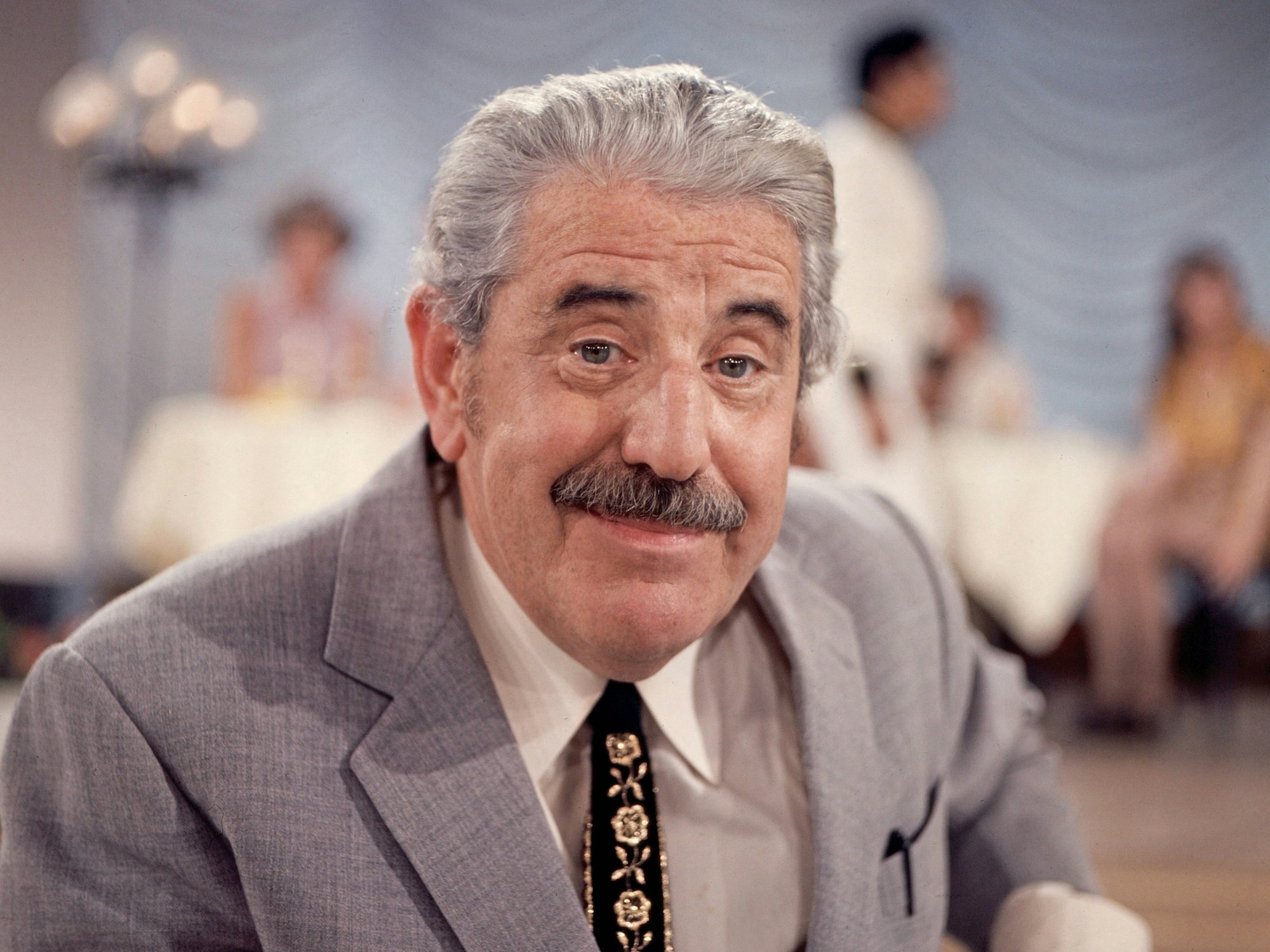 Willy Millowitsch, mit Anzug und Krawatte, lächelt in die Kamera.