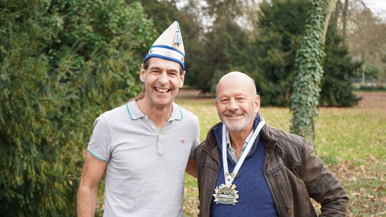 Zwei Männer stehen Arm in Arm in einem Park und strahlen in die Kamera.