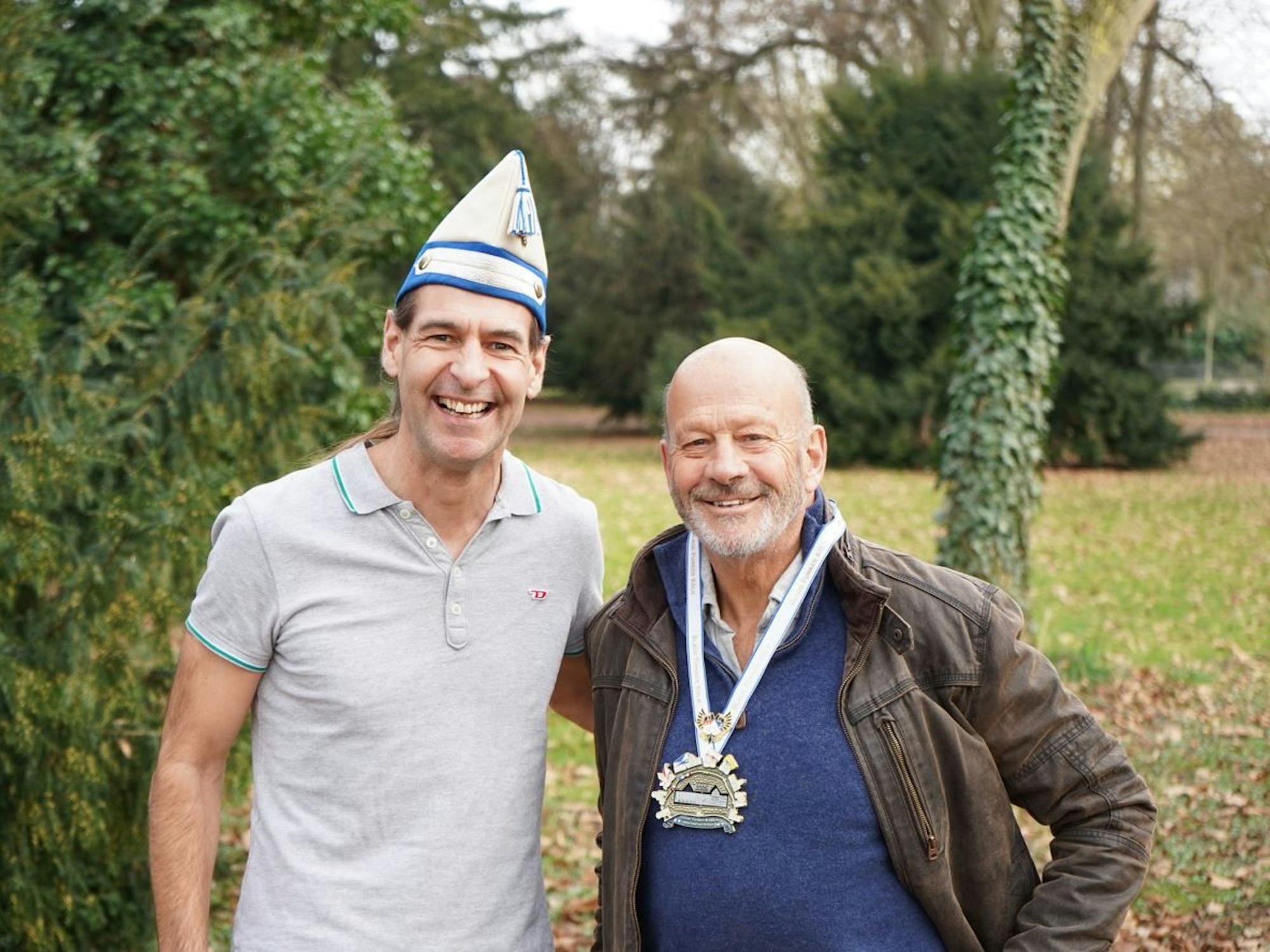 Zwei Männer stehen Arm in Arm in einem Park und strahlen in die Kamera.