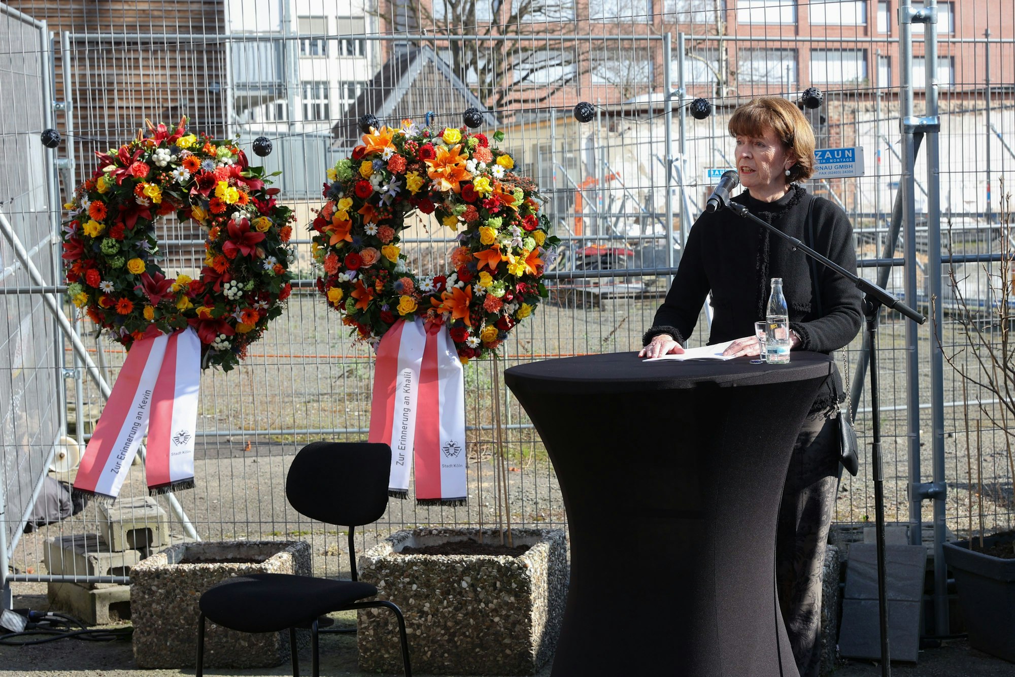 Oberbürgermeisterin Henriette Reker steht vor einem Mikrofon, hinter ihr an einem Bauzaun hängen zwei große Kränze mit Schleifen.