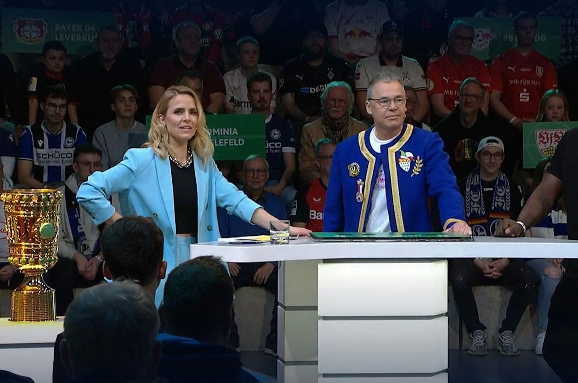 Andreas Rettig steht mit der Jacke des Kölner Festkomitees neben Moderatorin Stephanie Müller-Spirra.