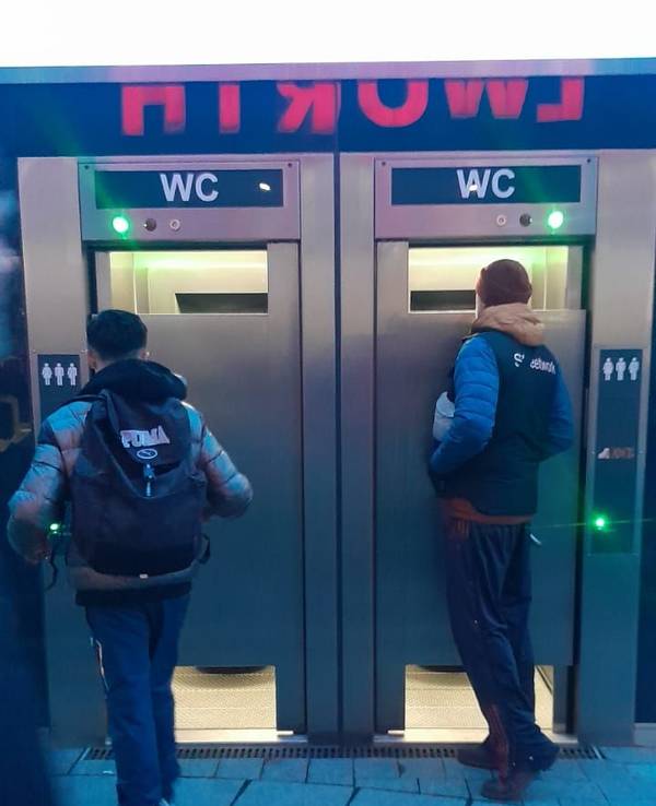 Zwei Männer stehen vor einer mobilen Toiletten und schauen durch große Spalte ins Innere.