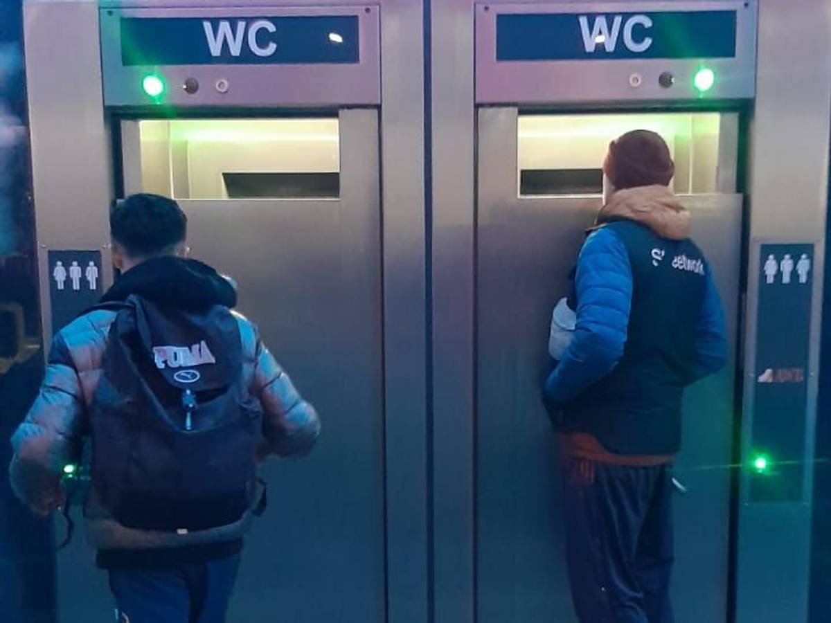 Zwei Männer stehen vor einer mobilen Toiletten und schauen durch große Spalte ins Innere.
