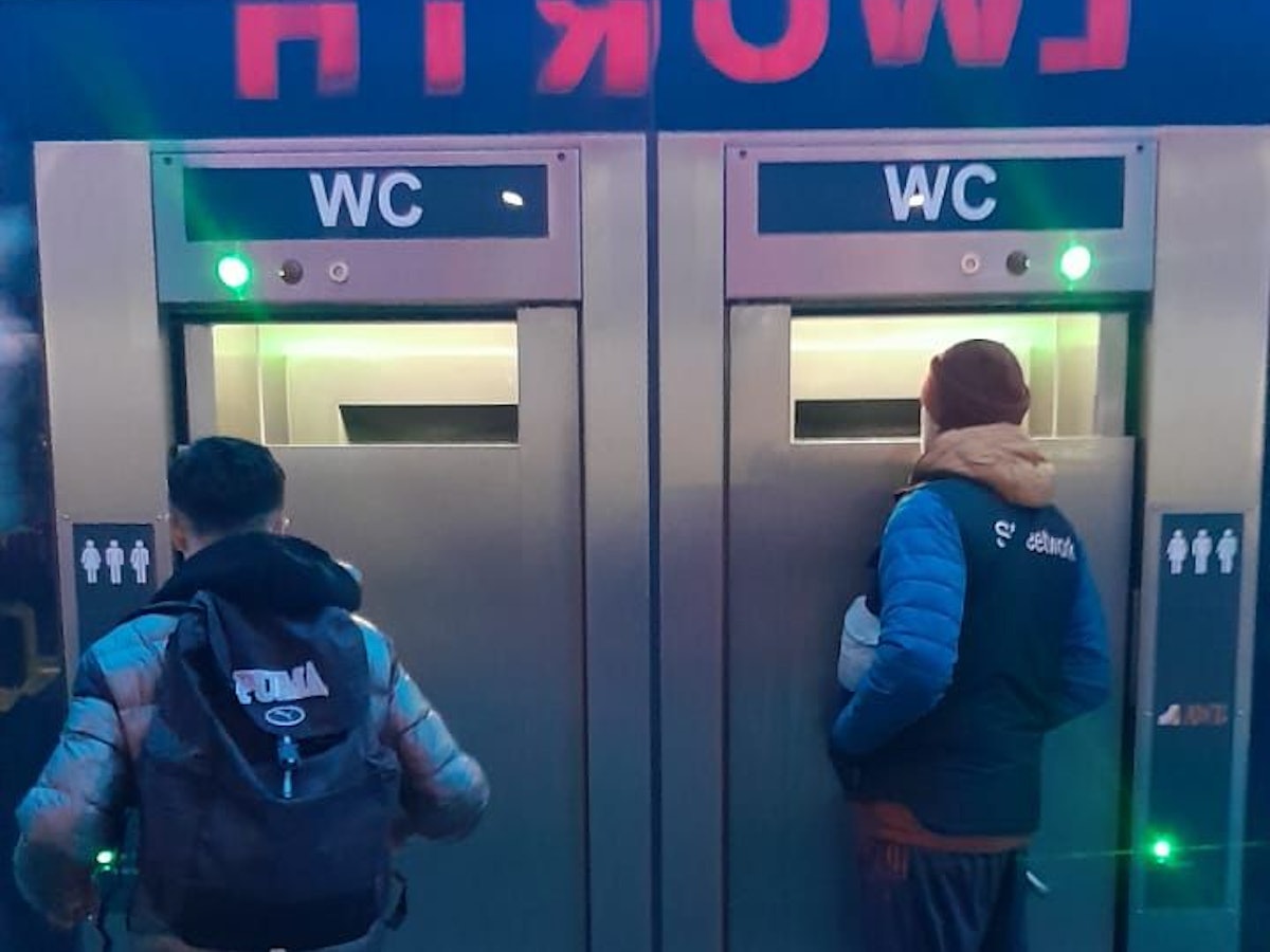 Zwei Männer stehen vor einer mobilen Toiletten und schauen durch große Spalte ins Innere.