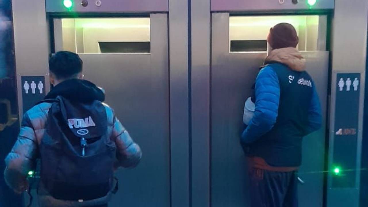 Zwei Männer stehen vor einer mobilen Toiletten und schauen durch große Spalte ins Innere.