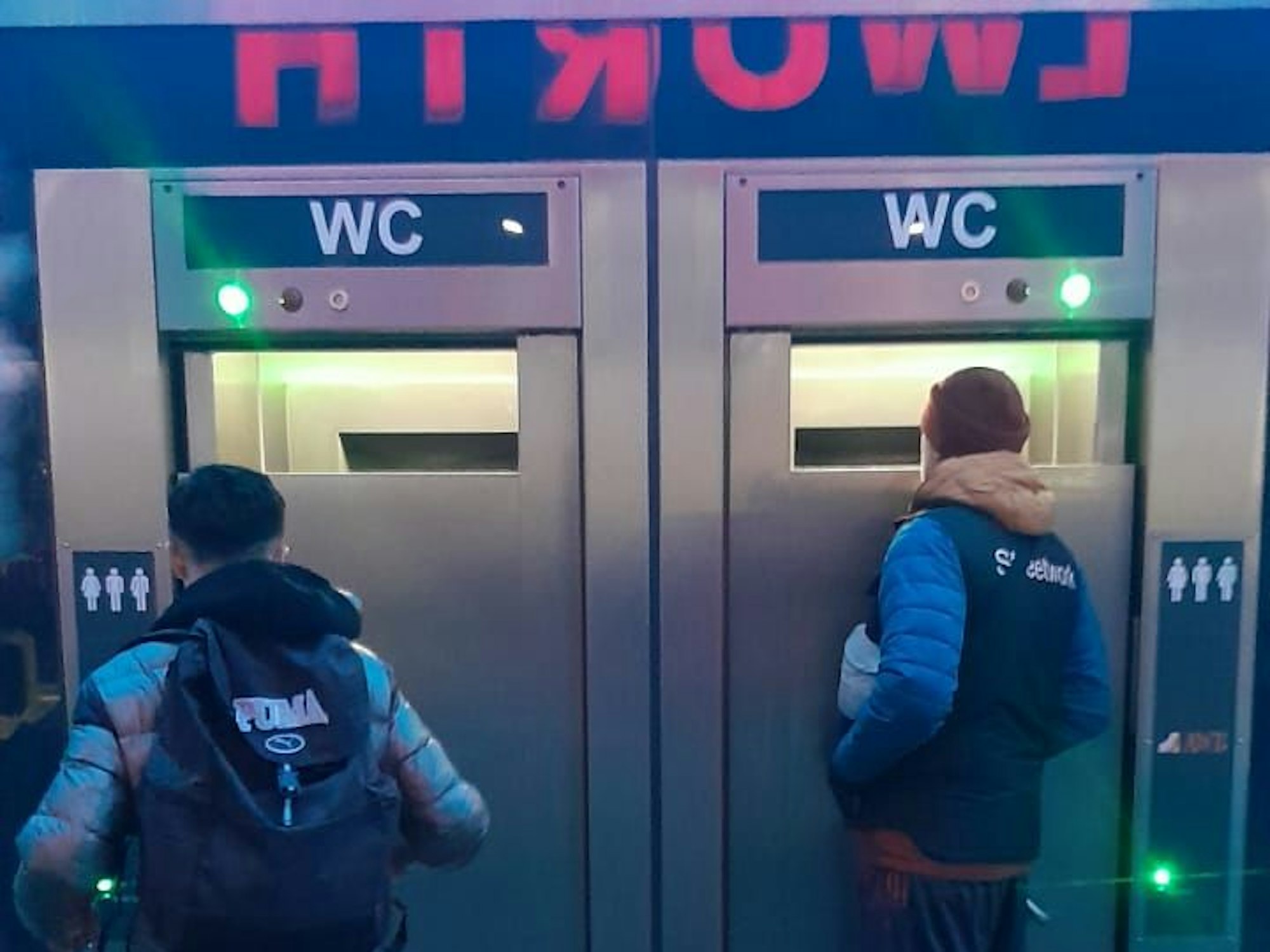 Zwei Männer stehen vor einer mobilen Toiletten und schauen durch große Spalte ins Innere.