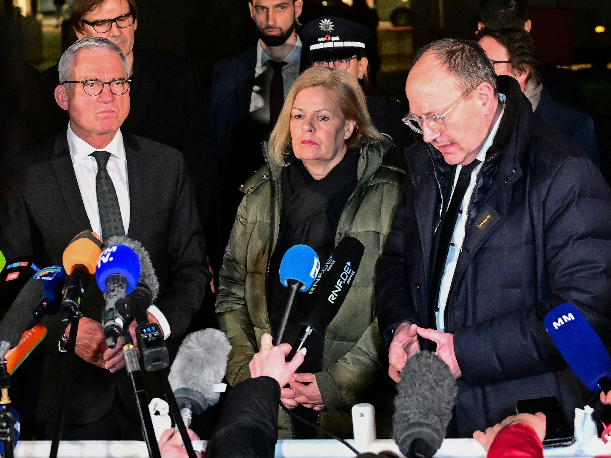 Zwei Männer und eine Frau vor Mikrofonen bei einem öffentlichen Statement.