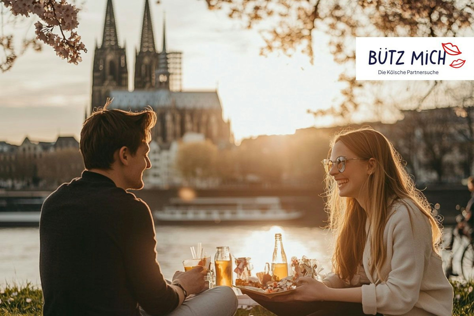 Ein Paar, das bei einem romantischen Frühlings-Picknick am Rheinufer in Köln sitzt, mit dem Kölner Dom im Hintergrund, während die Abendsonne warmes Licht auf die Szene wirft. Die beiden genießen Essen und Getränke, lachen miteinander und sind umgeben von blühenden Kirschbäumen, während sich Boote sanft auf dem Fluss bewegen.