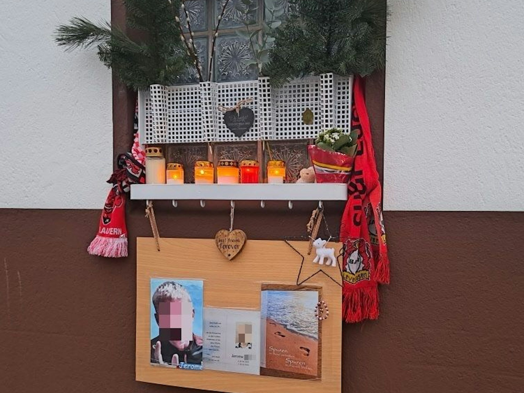 Eine Gedenktafel zeigt zwei Fotos des getöteten Jerome, darüber stehende brennende Kerzen, hängen zwei Fan-Schals unter anderem von Bayer Leverkusen.