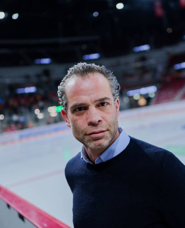 Niki Mondt steht vor der Eisfläche an der Bande und guckt in die Kamera.