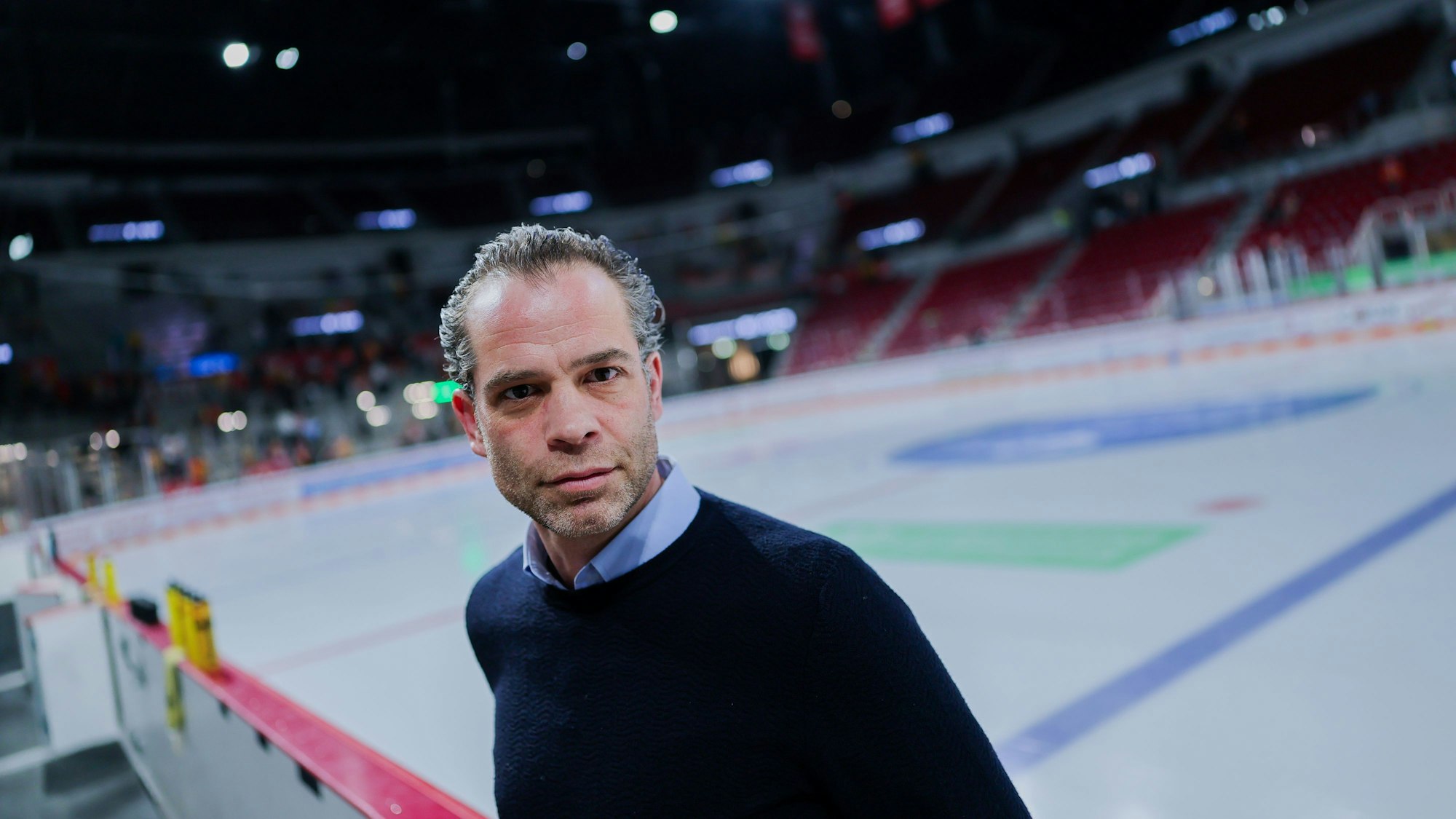Niki Mondt steht vor der Eisfläche an der Bande und guckt in die Kamera.