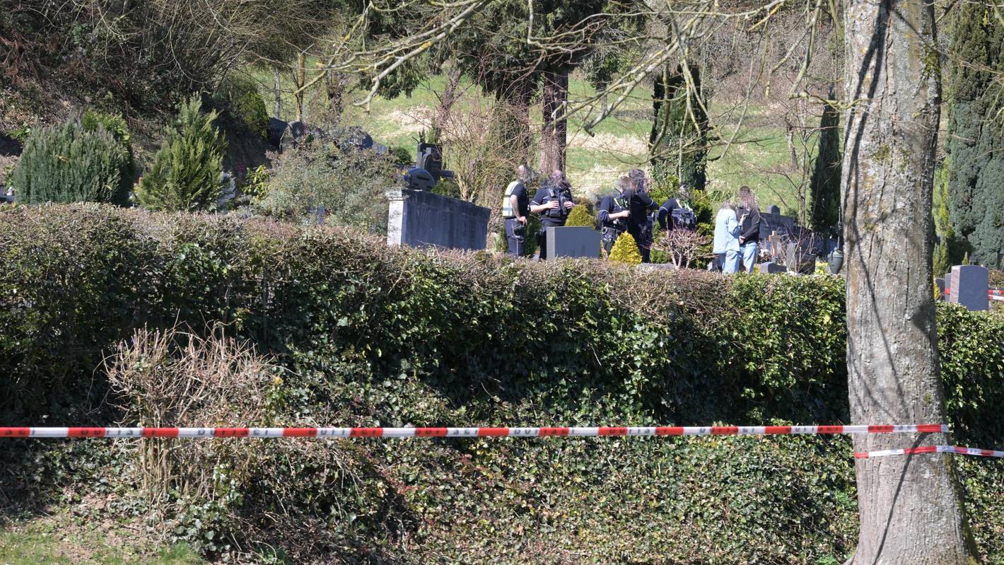 Ein Friedhof ist mit Polizei-Flatterband abgesperrt, zwischen Gräbern stehen unter anderem Polizeikräfte.